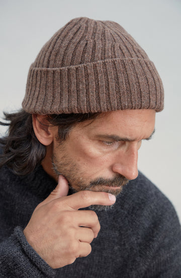 A close-up of a man wearing a brown wool ribbed knit beanie and a dark gray knit sweater, looking down thoughtfully against a light gray background. Made by Paul James Knitwear.