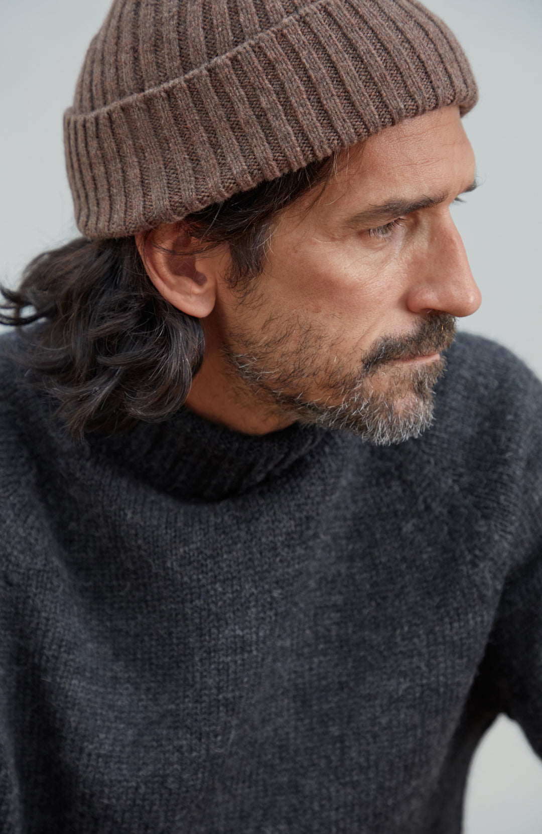 A close-up side profile of a man wearing a brown ribbed wool knit beanie and a dark charcoal gray knit sweater against a light gray background. Made by Paul James Knitwear. 