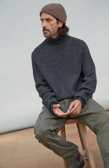 A man wearing a dark charcoal gray knit turtleneck wool sweater, olive green pants, and a brown ribbed wool beanie sits on a wooden stool against a light gray studio background. Made by Paul James Knitwear.