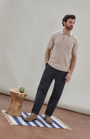 A man wearing a beige short-sleeve cable fisherman knit polo shirt by paul james knitwear with vertical cable and diamond patterns, paired with dark navy pants, against a solid light gray background.