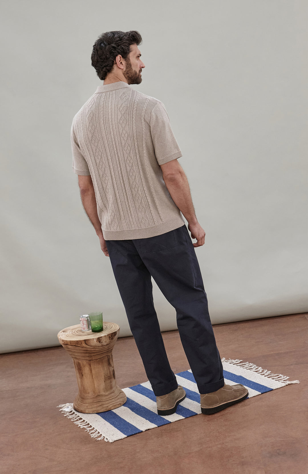 A man wearing a beige short-sleeve cable fisherman knit polo shirt by paul james knitwear with vertical cable and diamond patterns, paired with dark navy pants, against a solid light gray background.