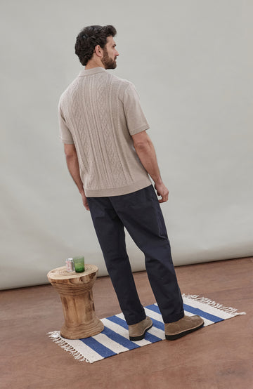 A man wearing a beige short-sleeve cable fisherman knit polo shirt by paul james knitwear with vertical cable and diamond patterns, paired with dark navy pants, against a solid light gray background.