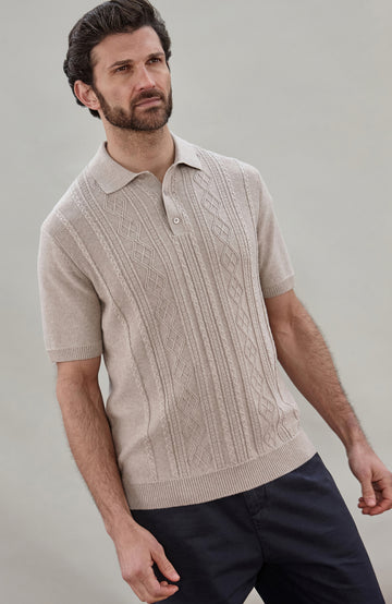 A man wearing a beige short-sleeve cable fisherman knit polo shirt by paul james knitwear with vertical cable and diamond patterns, paired with dark navy pants, against a solid light gray background.