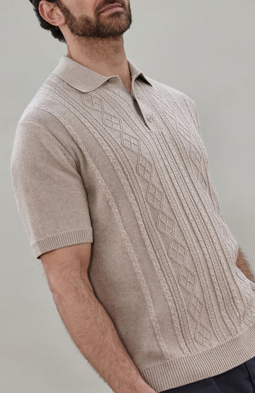 A man wearing a beige short-sleeve cable fisherman knit polo shirt by paul james knitwear with vertical cable and diamond patterns, paired with dark navy pants, against a solid light gray background.