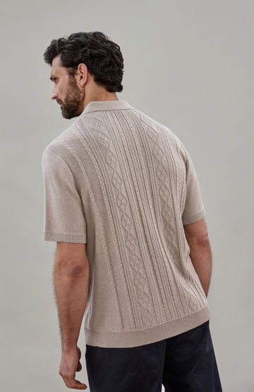 A man wearing a beige short-sleeve cable fisherman knit polo shirt by paul james knitwear with vertical cable and diamond patterns, paired with dark navy pants, against a solid light gray background.
