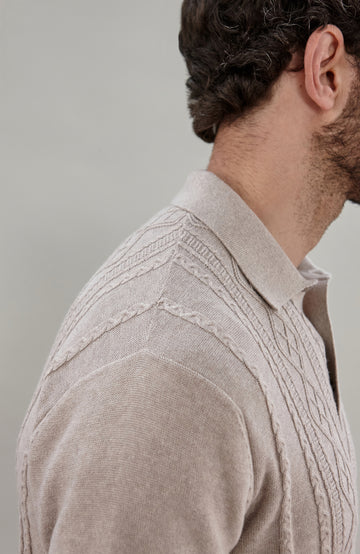 A man wearing a beige short-sleeve cable fisherman knit polo shirt by paul james knitwear with vertical cable and diamond patterns, paired with dark navy pants, against a solid light gray background.