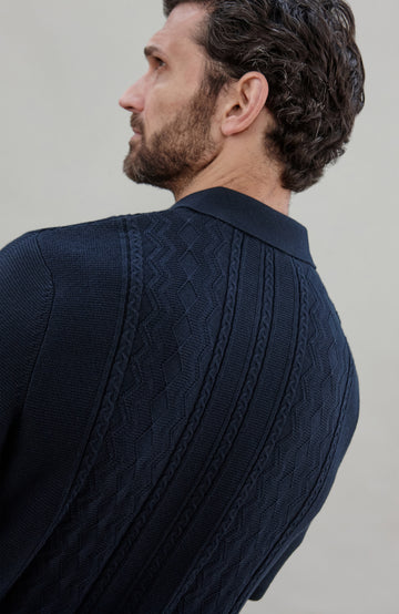 Man wearing a navy knit fisherman textured shirt by Paul James Knitwear and green pants holding a book against a plain background