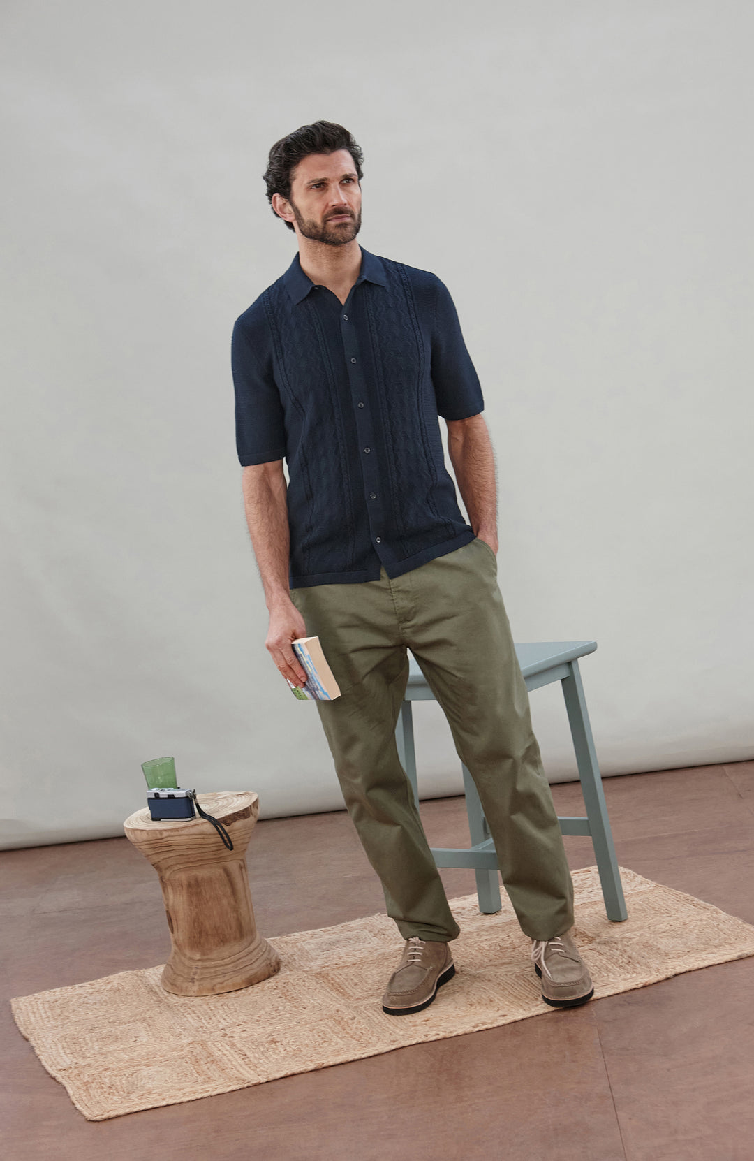Man wearing a navy knit fisherman textured shirt by Paul James Knitwear and green pants holding a book against a plain background