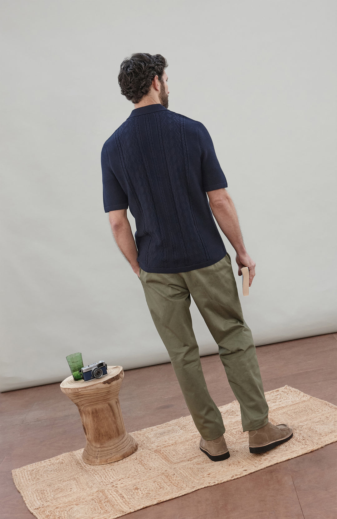 Man wearing a navy knit fisherman textured shirt by Paul James Knitwear and green pants holding a book against a plain background