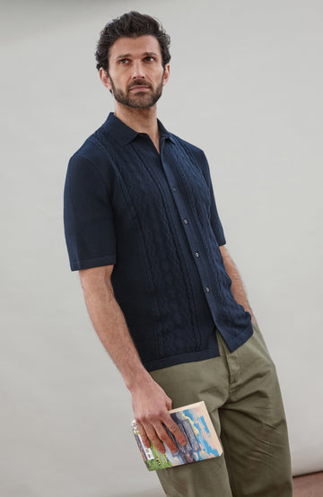 Man wearing a navy knit fisherman textured shirt by Paul James Knitwear and green pants holding a book against a plain background