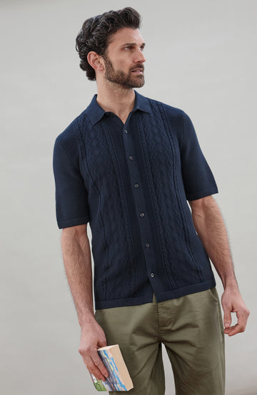 Man wearing a navy knit fisherman textured shirt by Paul James Knitwear and green pants holding a book against a plain background
