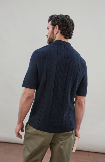 Man wearing a navy knit fisherman textured shirt by Paul James Knitwear and green pants holding a book against a plain background