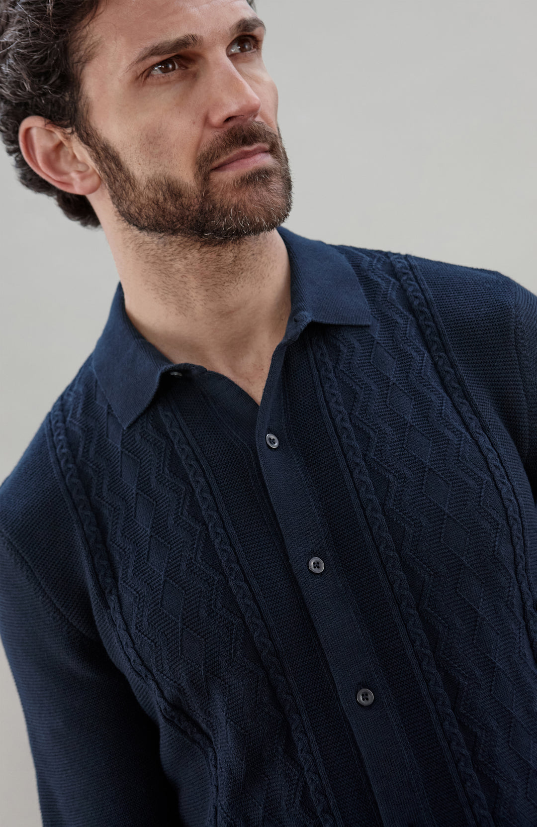 Man wearing a navy knit fisherman textured shirt by Paul James Knitwear and green pants holding a book against a plain background