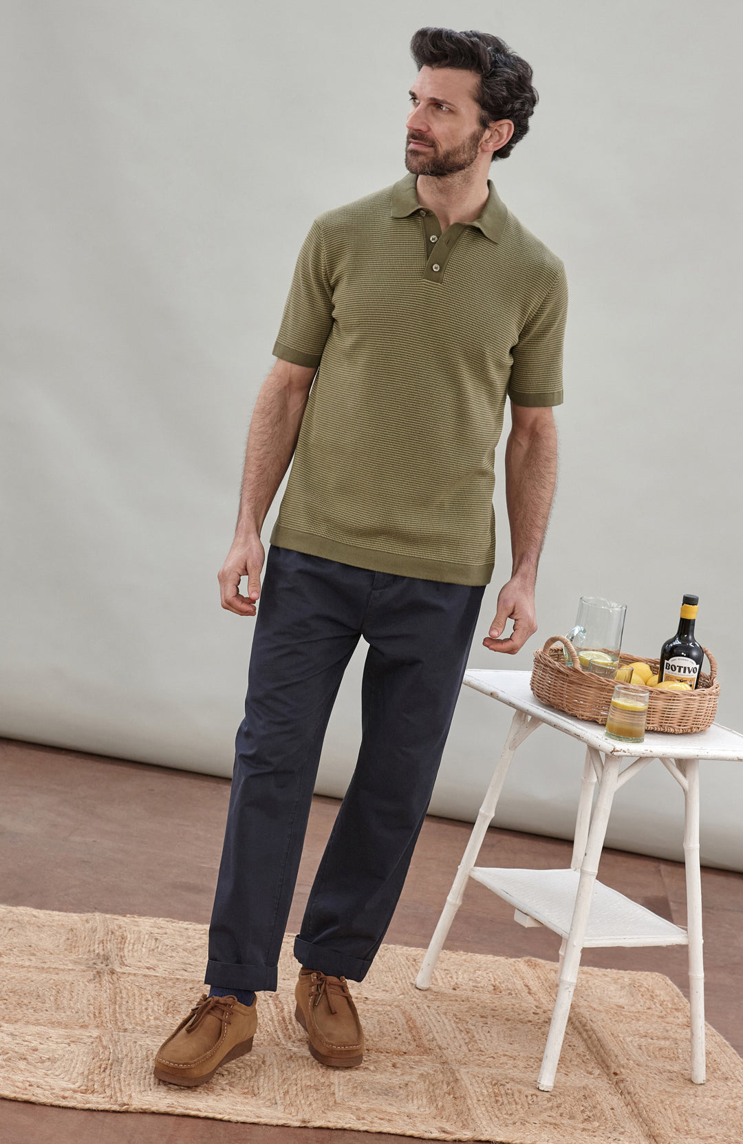 A man wearing a short-sleeve polo shirt bu Paul James Knitwear in olive green with fine horizontal stripes, paired with dark trousers, photographed against a plain light gray studio background.