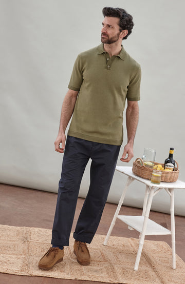 A man wearing a short-sleeve polo shirt bu Paul James Knitwear in olive green with fine horizontal stripes, paired with dark trousers, photographed against a plain light gray studio background.