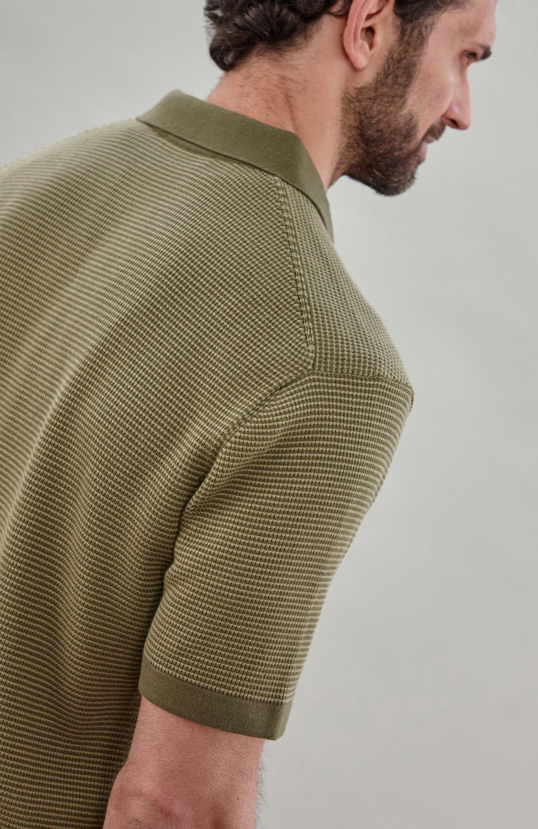 A man wearing a short-sleeve polo shirt bu Paul James Knitwear in olive green with fine horizontal stripes, paired with dark trousers, photographed against a plain light gray studio background.