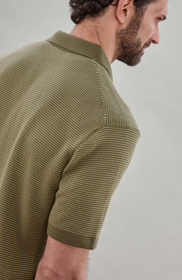 A man wearing a short-sleeve polo shirt bu Paul James Knitwear in olive green with fine horizontal stripes, paired with dark trousers, photographed against a plain light gray studio background.