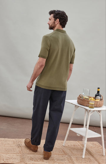 A man wearing a short-sleeve polo shirt bu Paul James Knitwear in olive green with fine horizontal stripes, paired with dark trousers, photographed against a plain light gray studio background.
