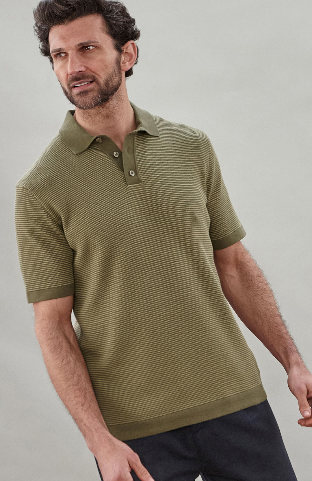 A man wearing a short-sleeve polo shirt bu Paul James Knitwear in olive green with fine horizontal stripes, paired with dark trousers, photographed against a plain light gray studio background.