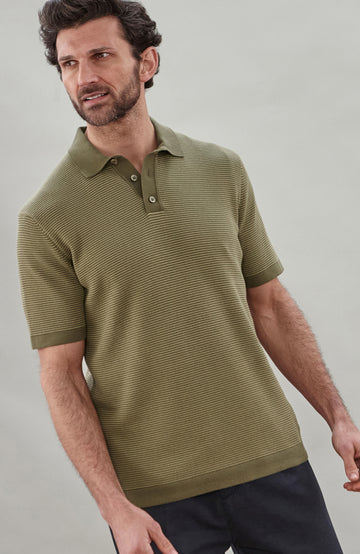 A man wearing a short-sleeve polo shirt bu Paul James Knitwear in olive green with fine horizontal stripes, paired with dark trousers, photographed against a plain light gray studio background.