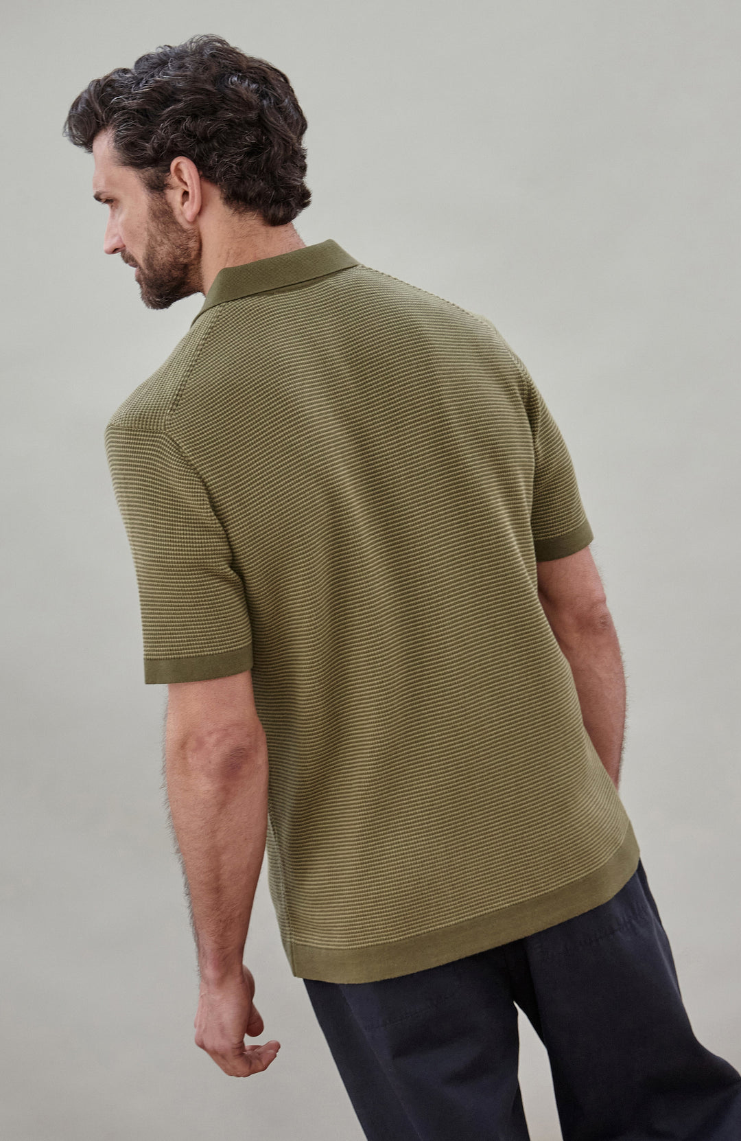 A man wearing a short-sleeve polo shirt bu Paul James Knitwear in olive green with fine horizontal stripes, paired with dark trousers, photographed against a plain light gray studio background.