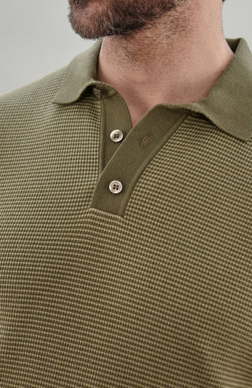 A man wearing a short-sleeve polo shirt bu Paul James Knitwear in olive green with fine horizontal stripes, paired with dark trousers, photographed against a plain light gray studio background.