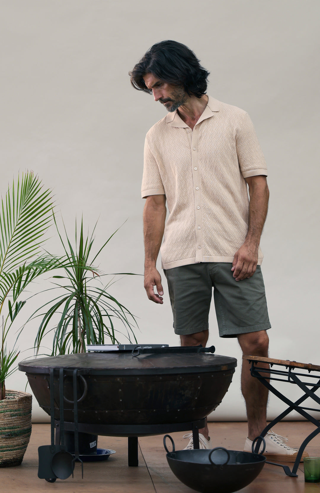 A man wearing a light beige short-sleeve button-up shirt and dark gray shorts stands next to a black round fire pit grill in an outdoor patio setting with potted. Made by Paul James Knitwear in cotton linen. plants.