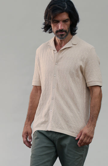 A man wearing a short-sleeve textured knit polo shirt in beige and olive green pants, standing against a neutral gray studio background. Made by Paul James Knitwear in cotton linen.