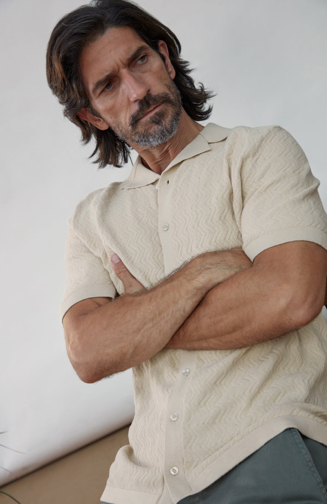 A man with dark hair and a beard wearing a textured cream short-sleeve button-up shirt, posing with his arms crossed against a neutral light gray background. Made by Paul James Knitwear in cotton linen.