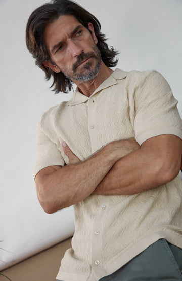 A man with dark hair and a beard wearing a textured cream short-sleeve button-up shirt, posing with his arms crossed against a neutral light gray background. Made by Paul James Knitwear in cotton linen.