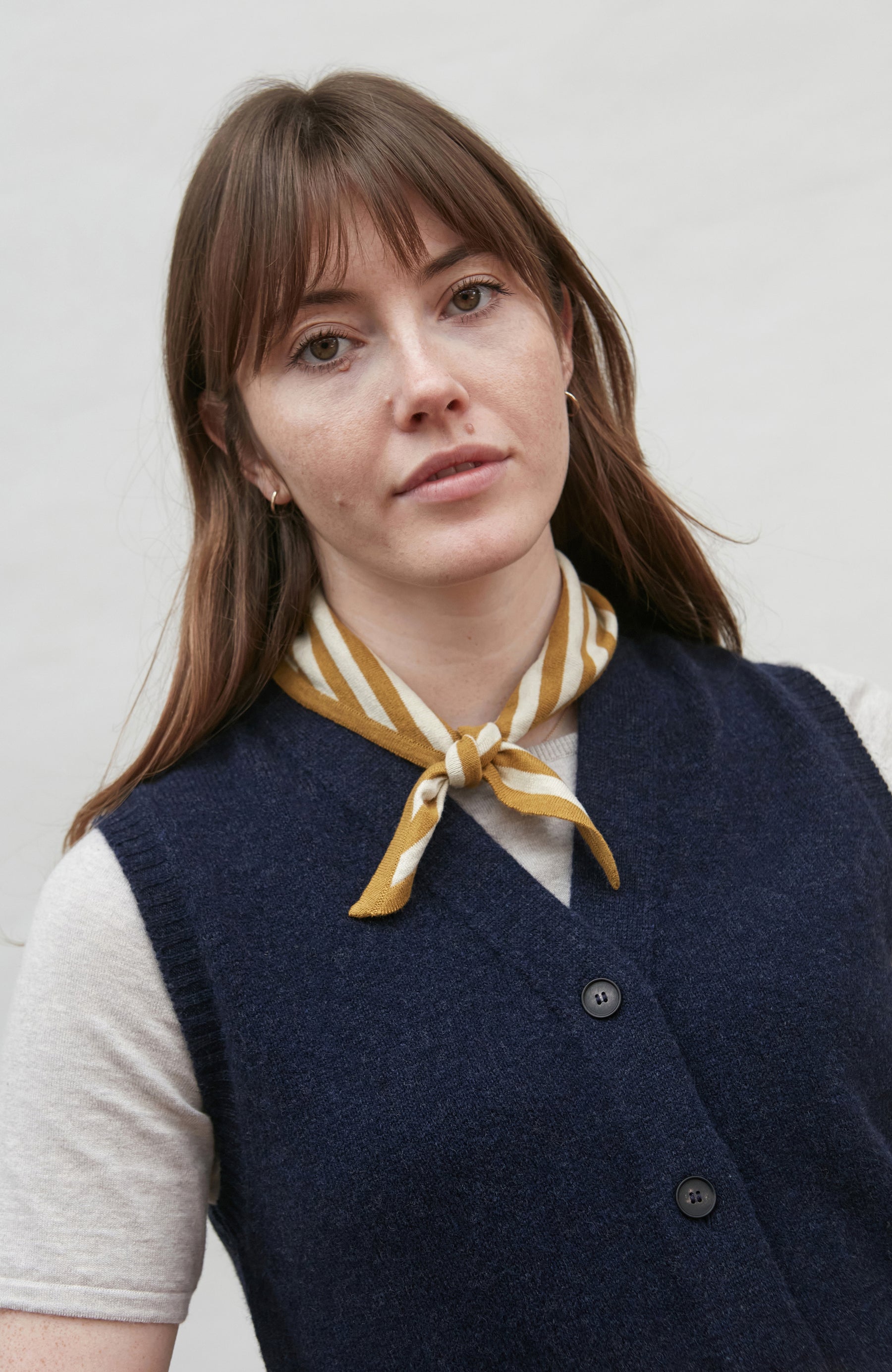 Woman wearing a navy wool vest and white shirt with a yellow stripe scarf, against a plain background