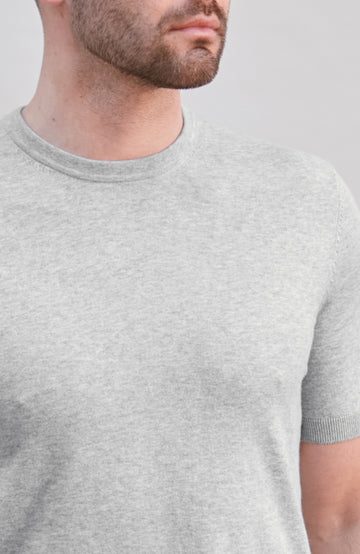 A close-up of a man wearing a light heather gray short-sleeve crewneck t-shirt against a plain light gray background.