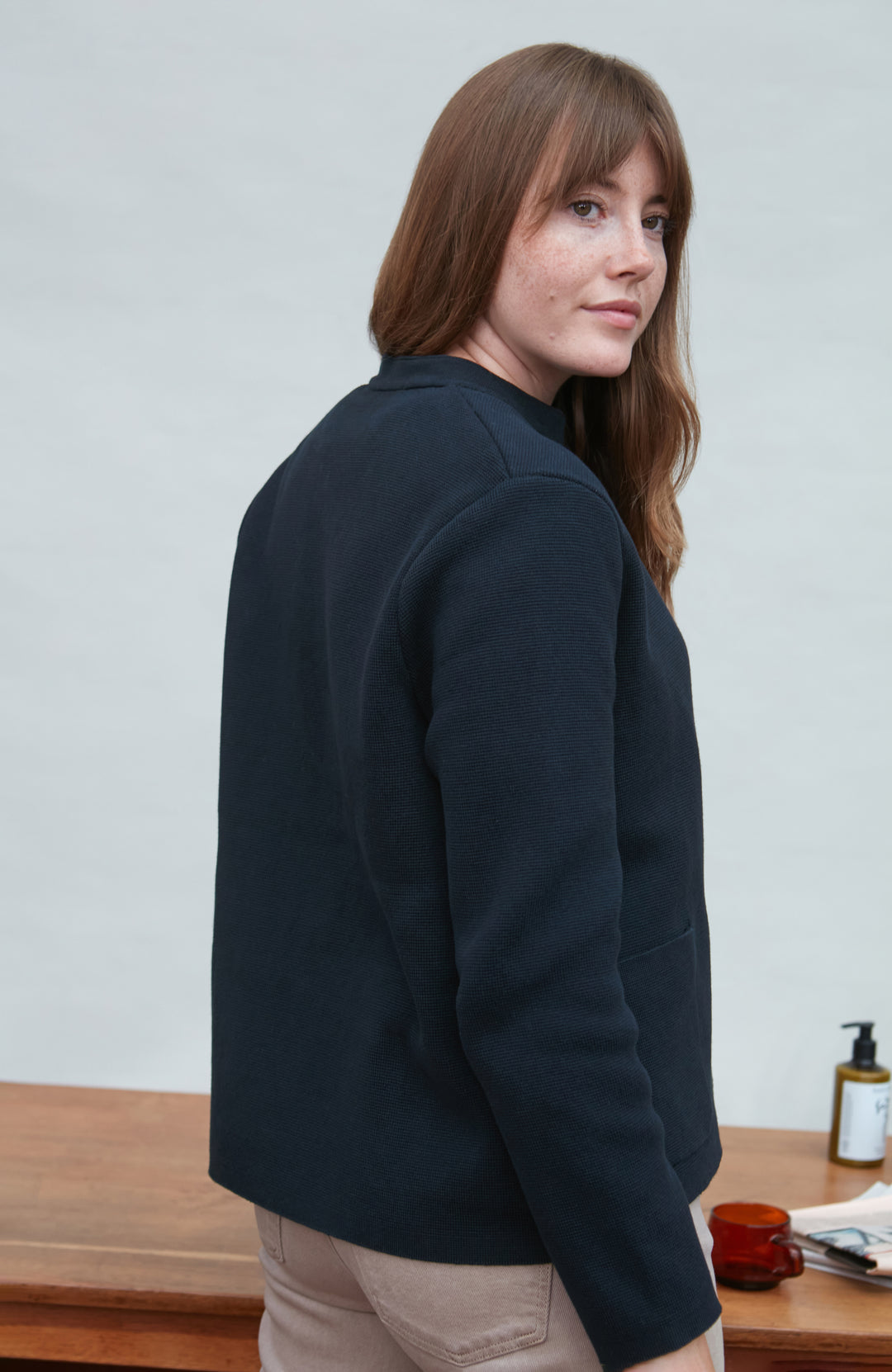 Woman wearing a navy blue jacket standing in front of a wooden table with a white wall background