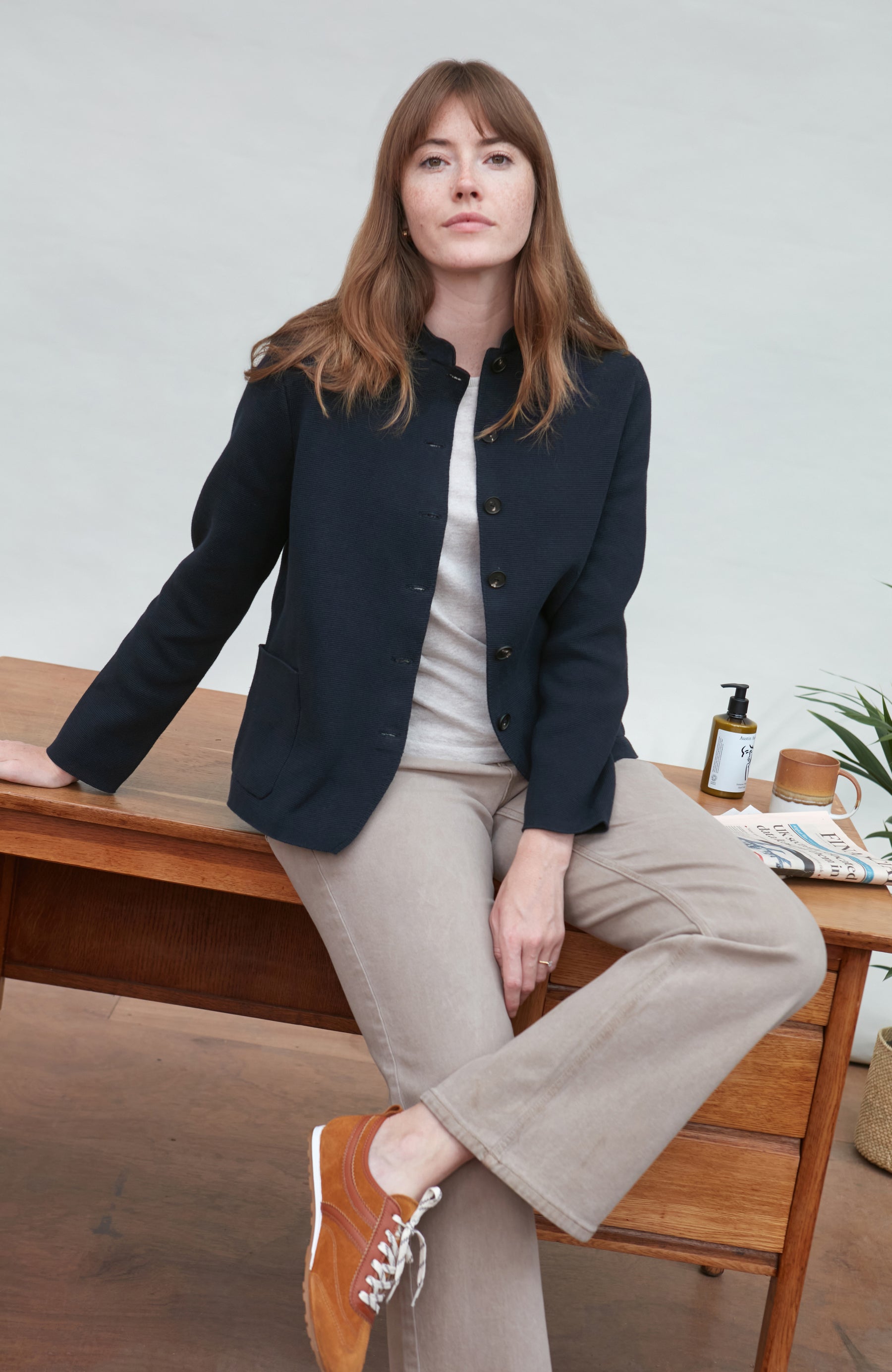 Woman sitting on a wooden desk wearing a navy nehru jacket by paul james and beige pants.