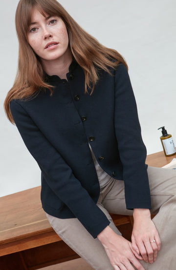 Woman wearing a navy paul james nehru jacket sitting on a wooden bench with a white background