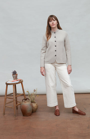 Woman wearing a light beige nehru jacket and white pants standing in a room with a wooden floor and white walls.