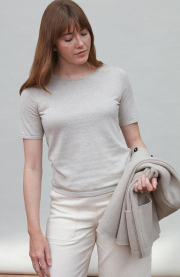 Woman wearing a light gray t-shirt By Paul James Knitwear and white pants holding a beige jacket against a plain background