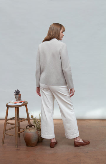 Woman wearing a light gray nehru jacket and white pants standing in a room with a wooden stool and plants.