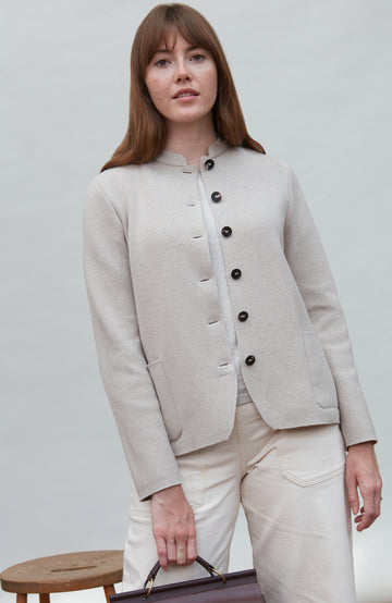 Woman wearing a beige paul james mandarin colllar jacket and pants holding a brown bag against a light gray background