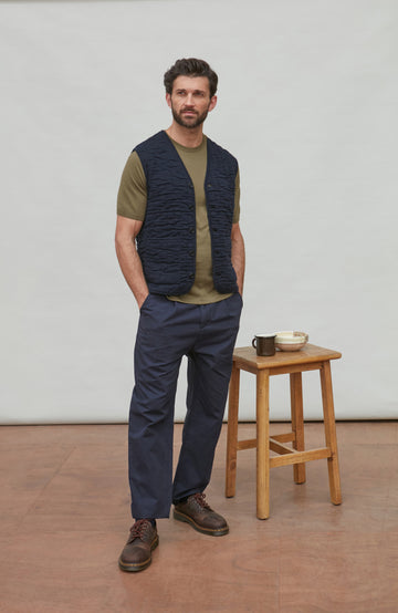 Man wearing Paul James gilet standing in a minimalistic room with a wooden stool and small items.