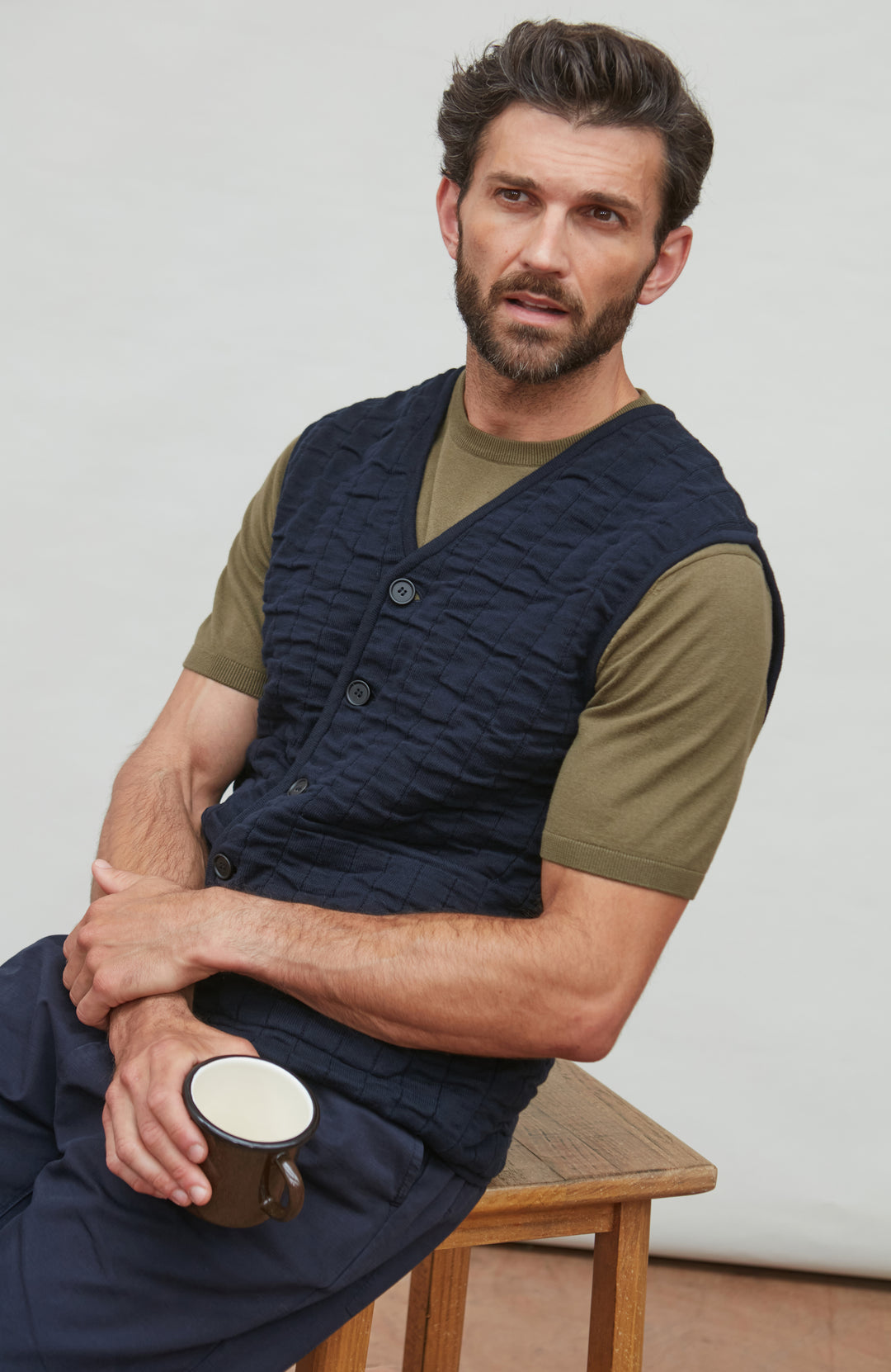 Man wearing a navy quitled vest by paul james over an olive green shirt, holding a mug.