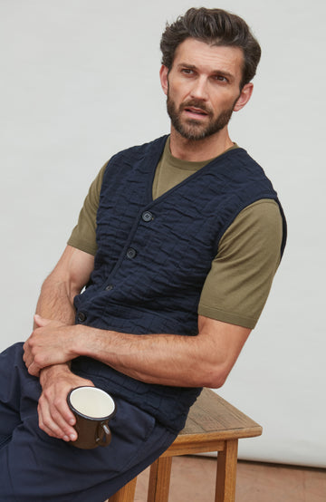 Man wearing a navy quitled vest by paul james over an olive green shirt, holding a mug.