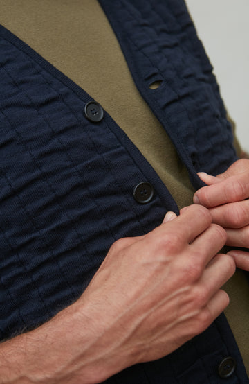 Close-up of a person buttoning a navy blue textured shirt against a beige background