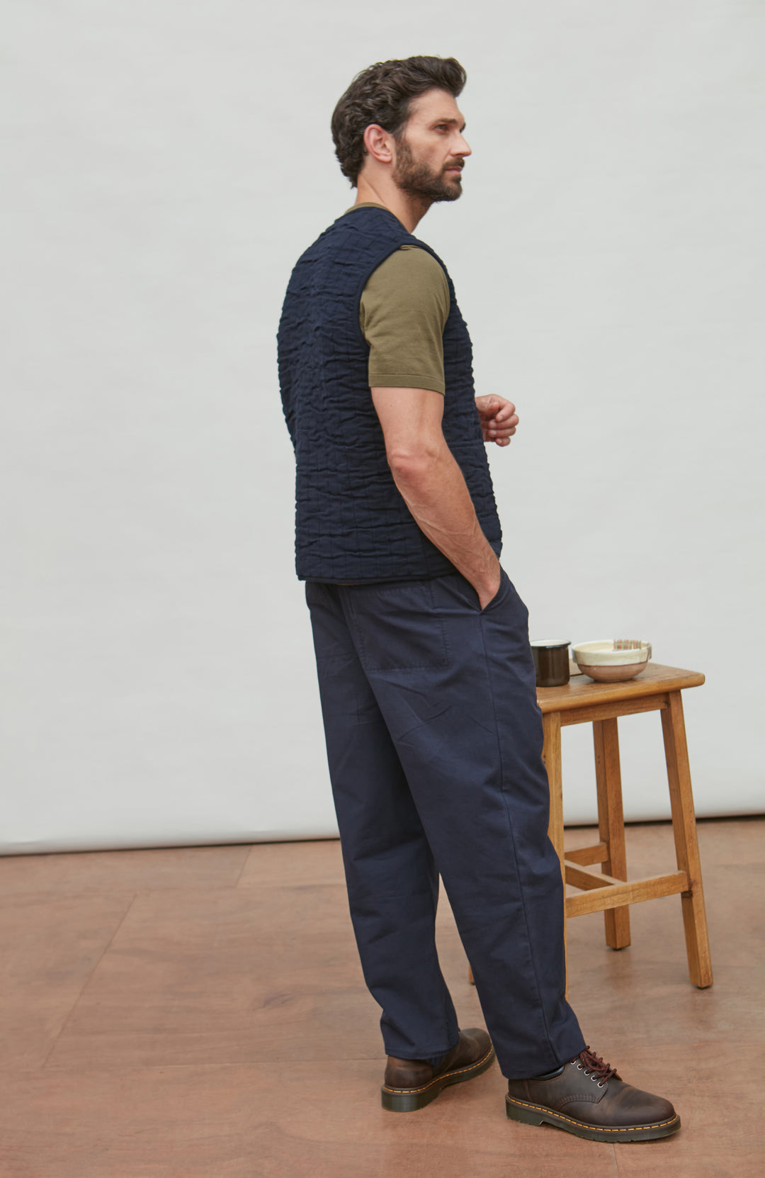 Man wearing a navy vest over a green shirt with dark pants, standing in a minimalistic room.