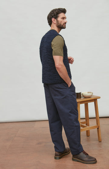 Man wearing a navy vest over a green shirt with dark pants, standing in a minimalistic room.