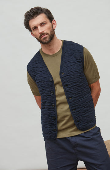 Man wearing a navy quilted vest over a green shirt with a neutral background