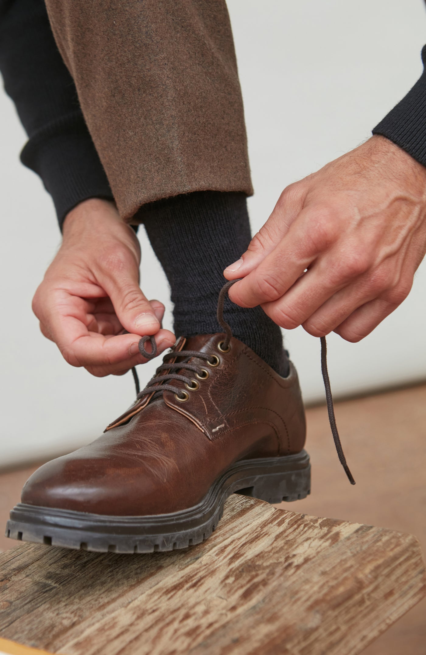 Person brown alpaca socks tying the laces of a brown leather shoe on a wooden floor.