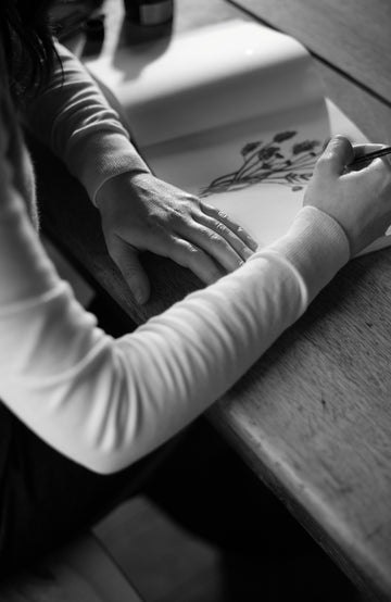 Person drawing on a piece of paper with a pen at a desk by Paul James Knitwear