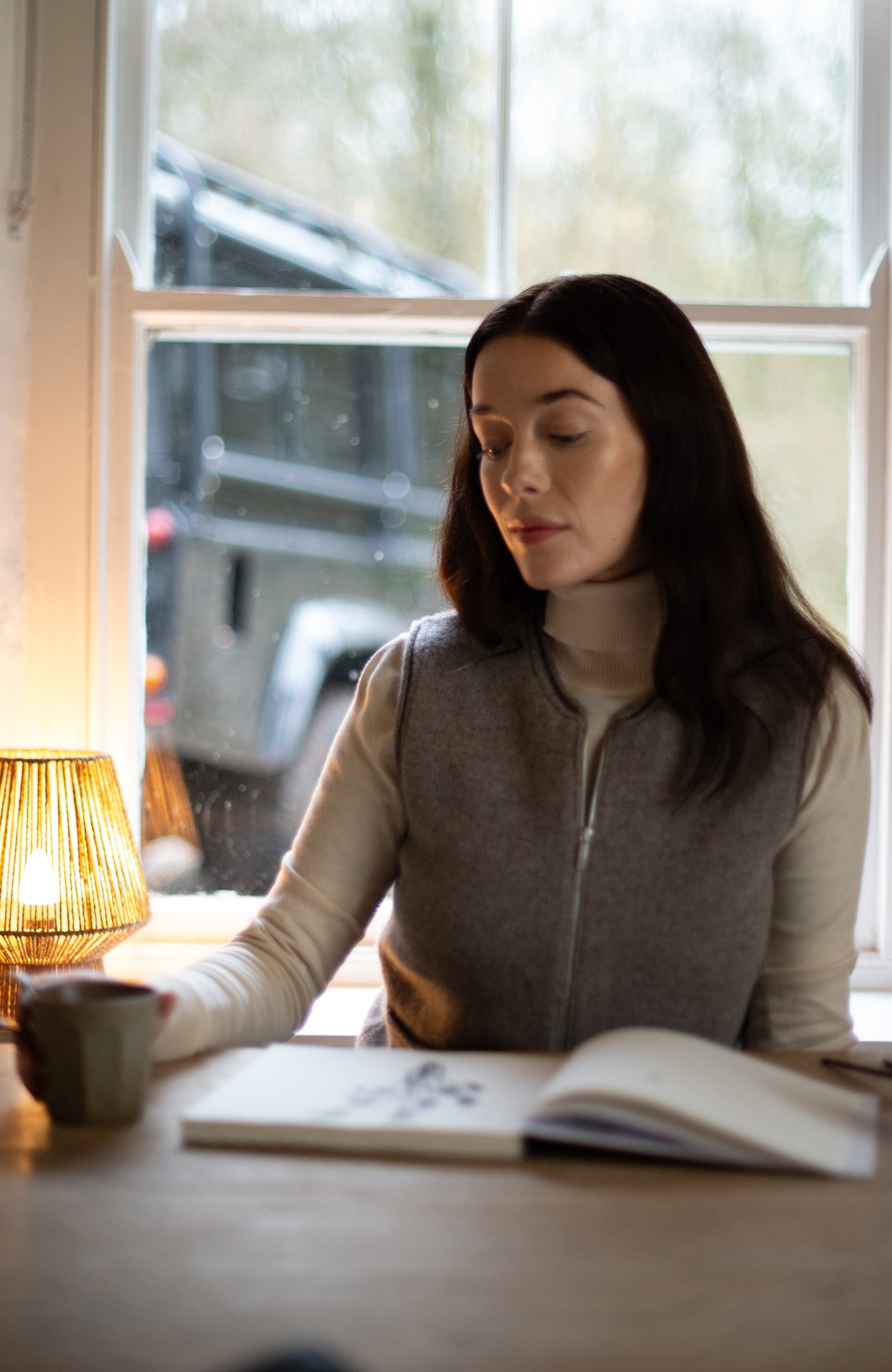 A woman with long dark hair sits at a wooden table by a window, wearing a gray zip-up wool vest by paul james knitwear over a cream turtleneck. An open notebook and a lit lamp are on the table in front of her.