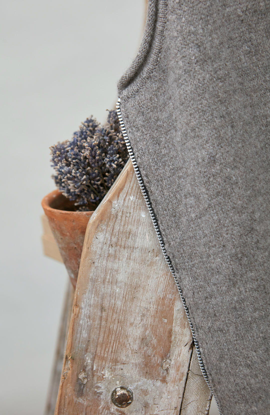 Gray sleeveless wool gilet by paul james with a wooden vase and lavender plant on a light background